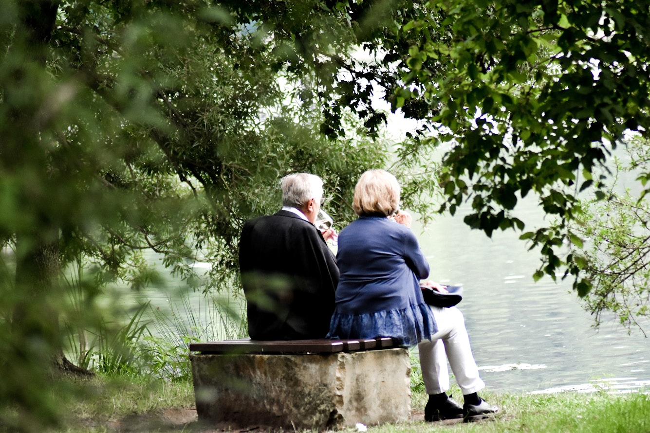 An einem See neben einem Wald, sitzt auf einer Parkbank ein Paar im Rentenalter und schauen auf den See. Altersvorsorge mit Gold und Silber als Vertriebsprodukt.